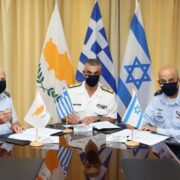 Tre oficerë ushtarakë nga Izraeli, Greqia dhe Qiproja gjatë një takimi bashkëpunimi në Greqi, me flamujt e tri vendeve në sfond.